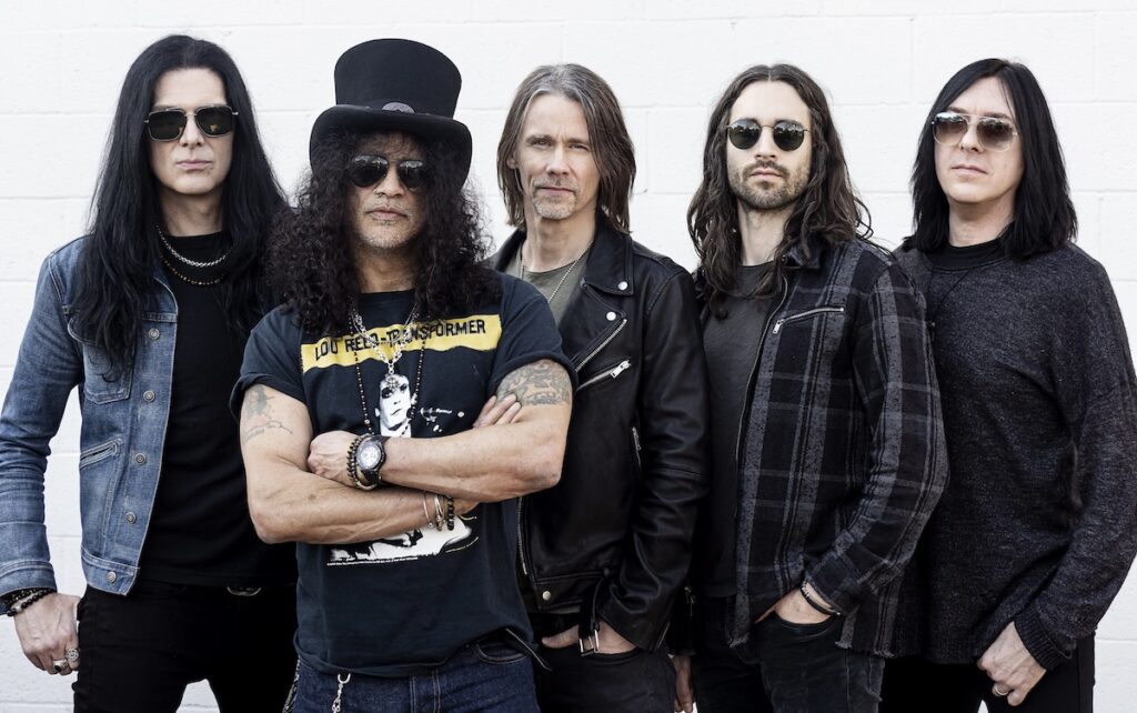 Band members standing in a row with Slash slightly in front in top hat, sunglasses and graphic T