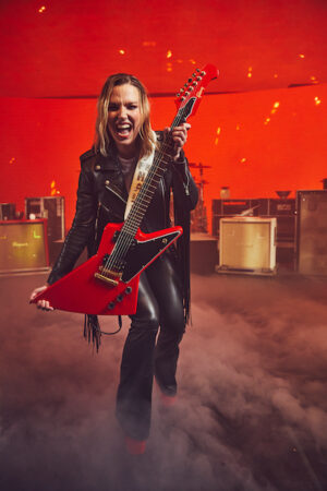 Singer-guitarist Lzzy Hale with her new namesake cardinal red Gibson Lzzy Hale Explorerbird held out in front of her. She wears a black leather jacket and leather pants in clouds of smoke in front of equipment on a stage.